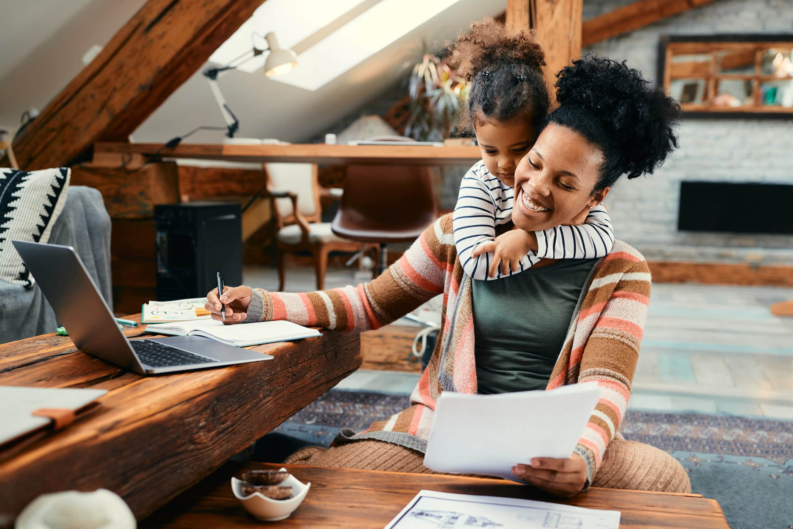 Small black girl embracing her busy mother who is working at home.