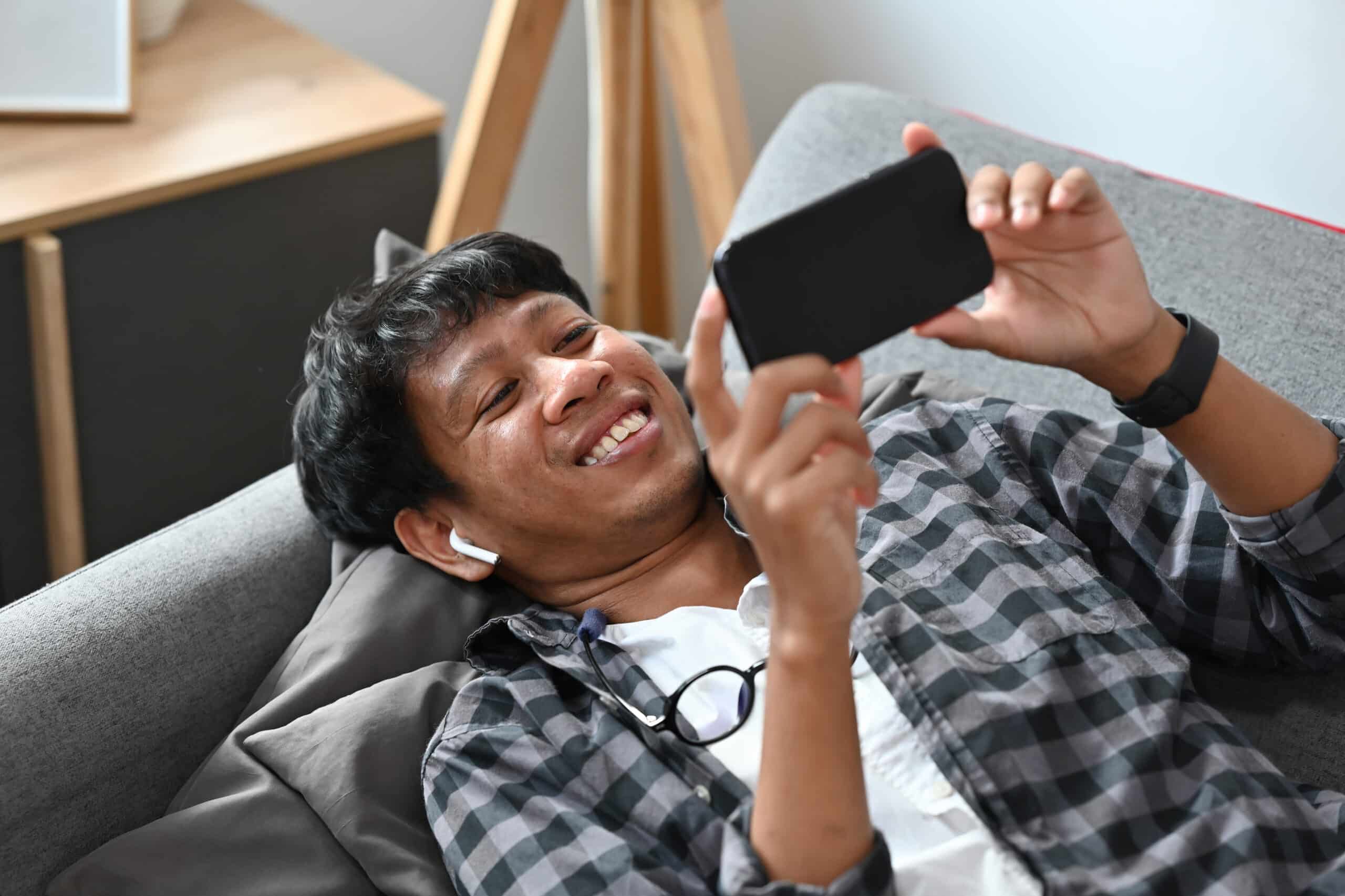 Asian man smiling with airpods in while taking surveys for money on his phone on the couch.
