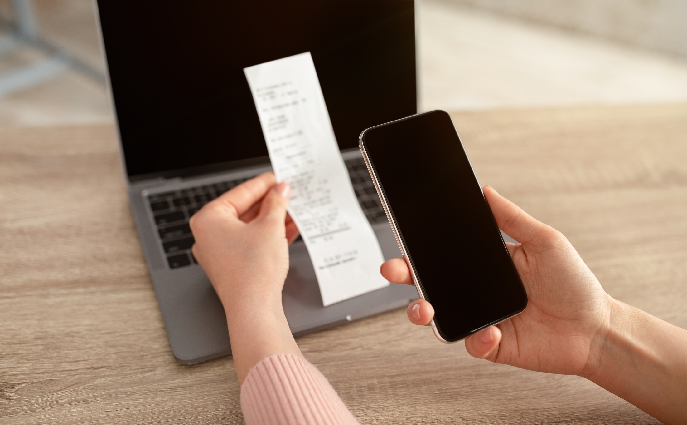 receipt app, woman's hand holding receipt and taking a picture of it with cell phone, in front of a computer.